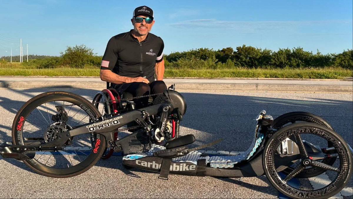 A man wearing a black cycling jersey, black cap, and sunglasses sits on a black trike with a rear cargo bed and a separate front wheel assembly, photographed outdoors on a paved road with trees and blue sky in the background. The bike frame and front wheel assembly show the brand name Earth Bike.